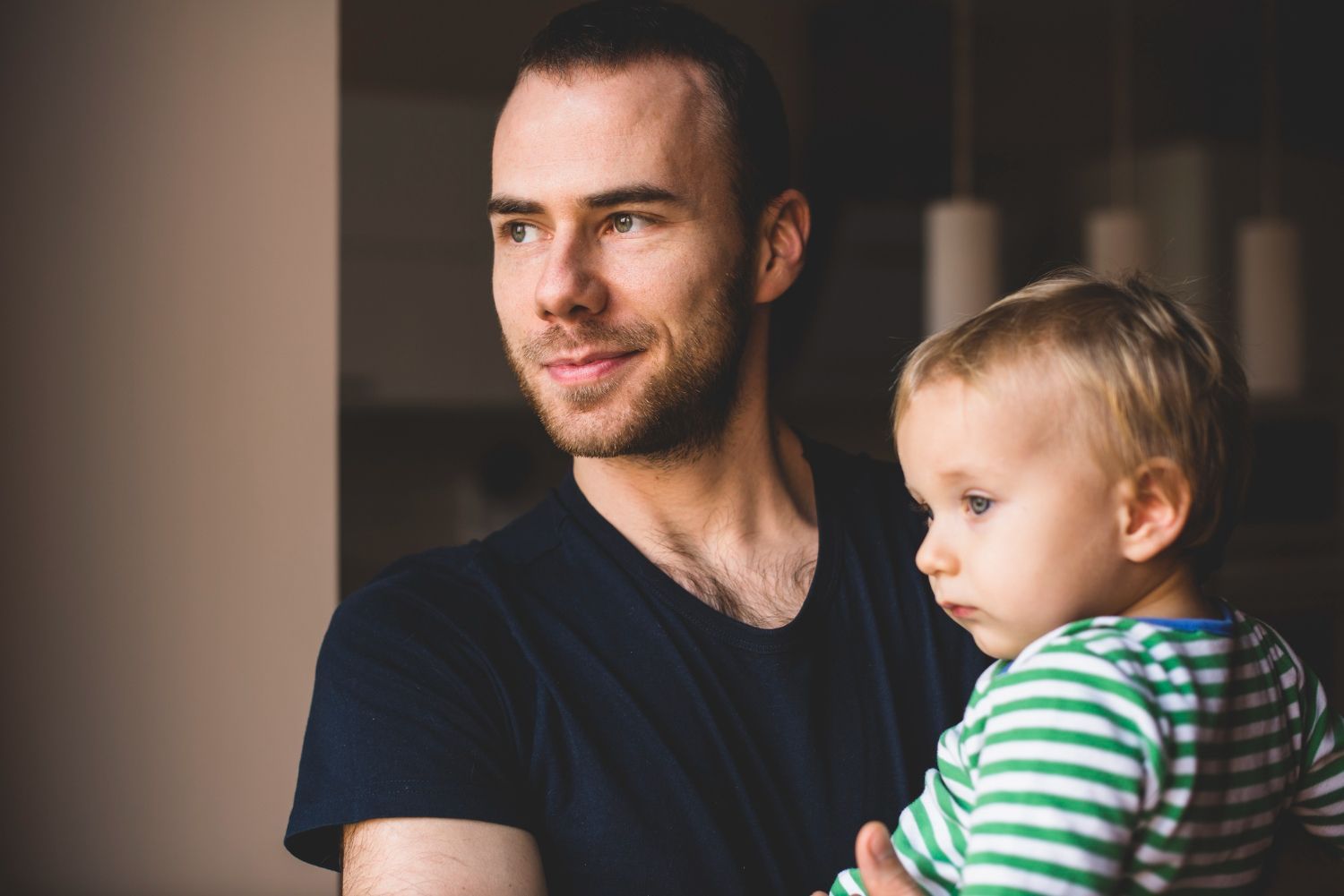 Man holding a child, both looking off-camera. The man has a small beard and wears a dark blue shirt.