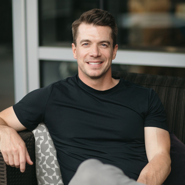 Man in black t-shirt smiles, sitting in outdoor chair.