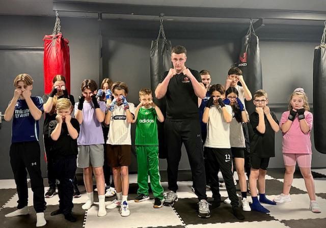 Een groep kinderen en een instructeur poseren in een bokszaal, met de bokshandschoenen in een vechtpositie.