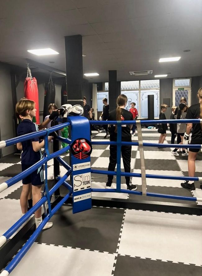 Mensen trainen in een boksring in een sportschool met blauwe touwen en zwart-witte vloermatten.