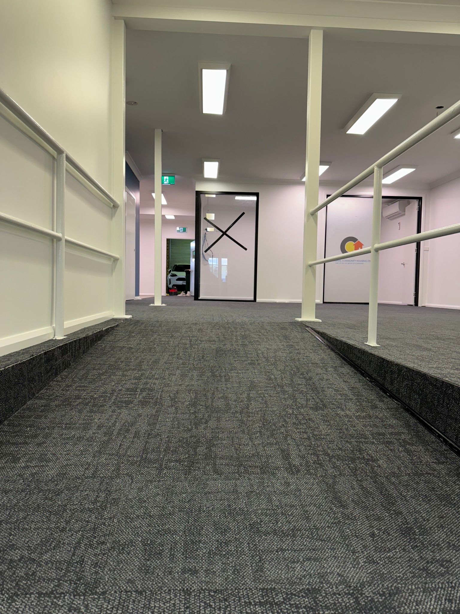 A ramp with grey patterned carpet and white railings leading to a hallway with glass-paned doors.