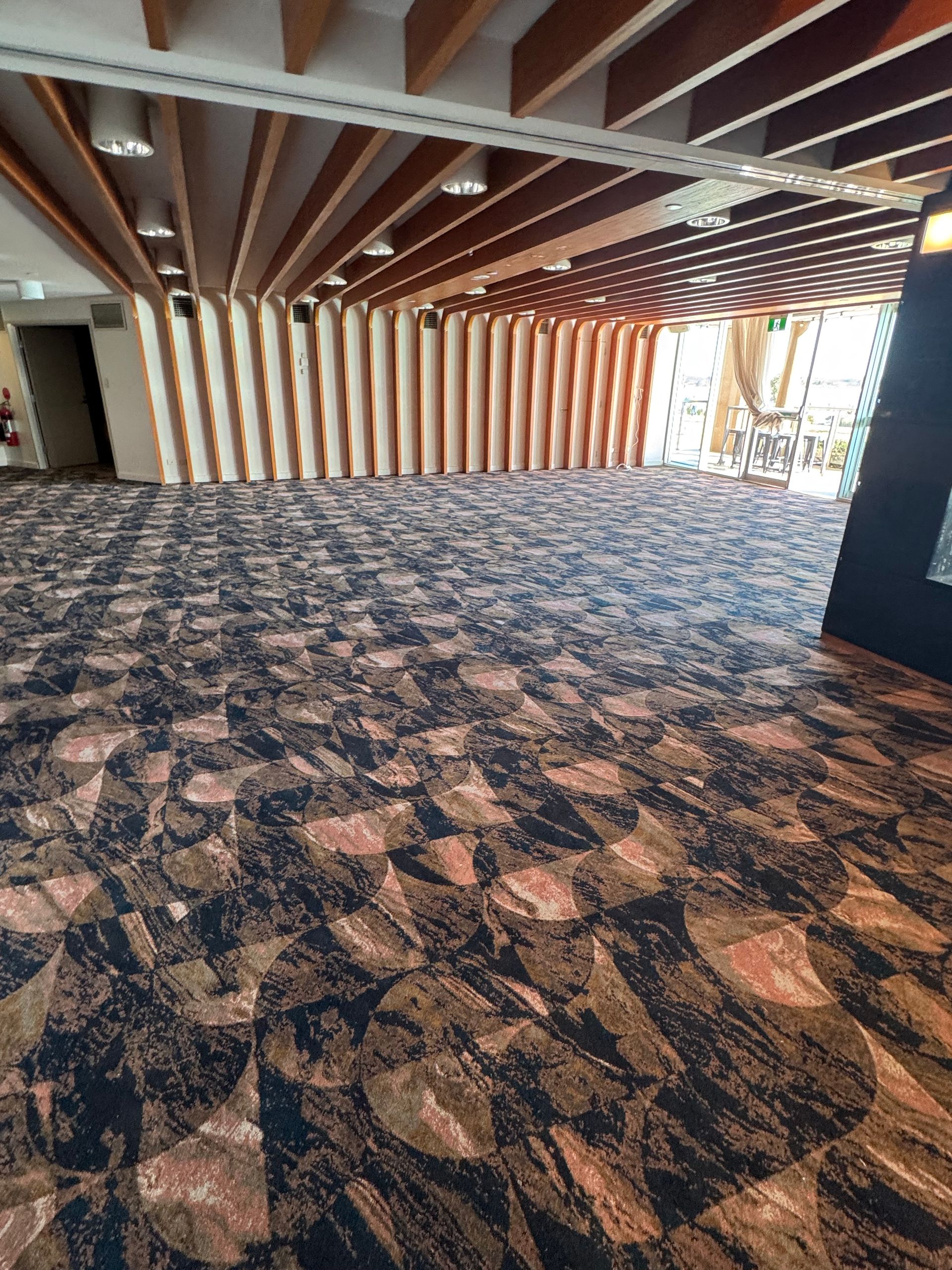 Empty carpeted room with exposed wooden ceiling beams and a window wall on the right.