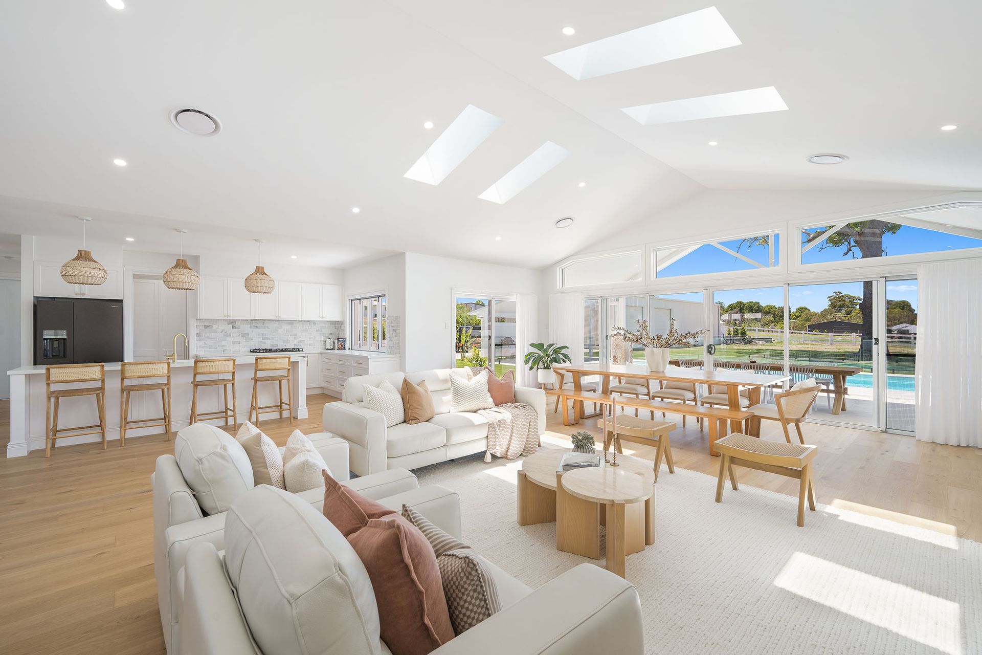 Bright open living room with white sofas, wooden furniture, and large windows overlooking a sunny landscape