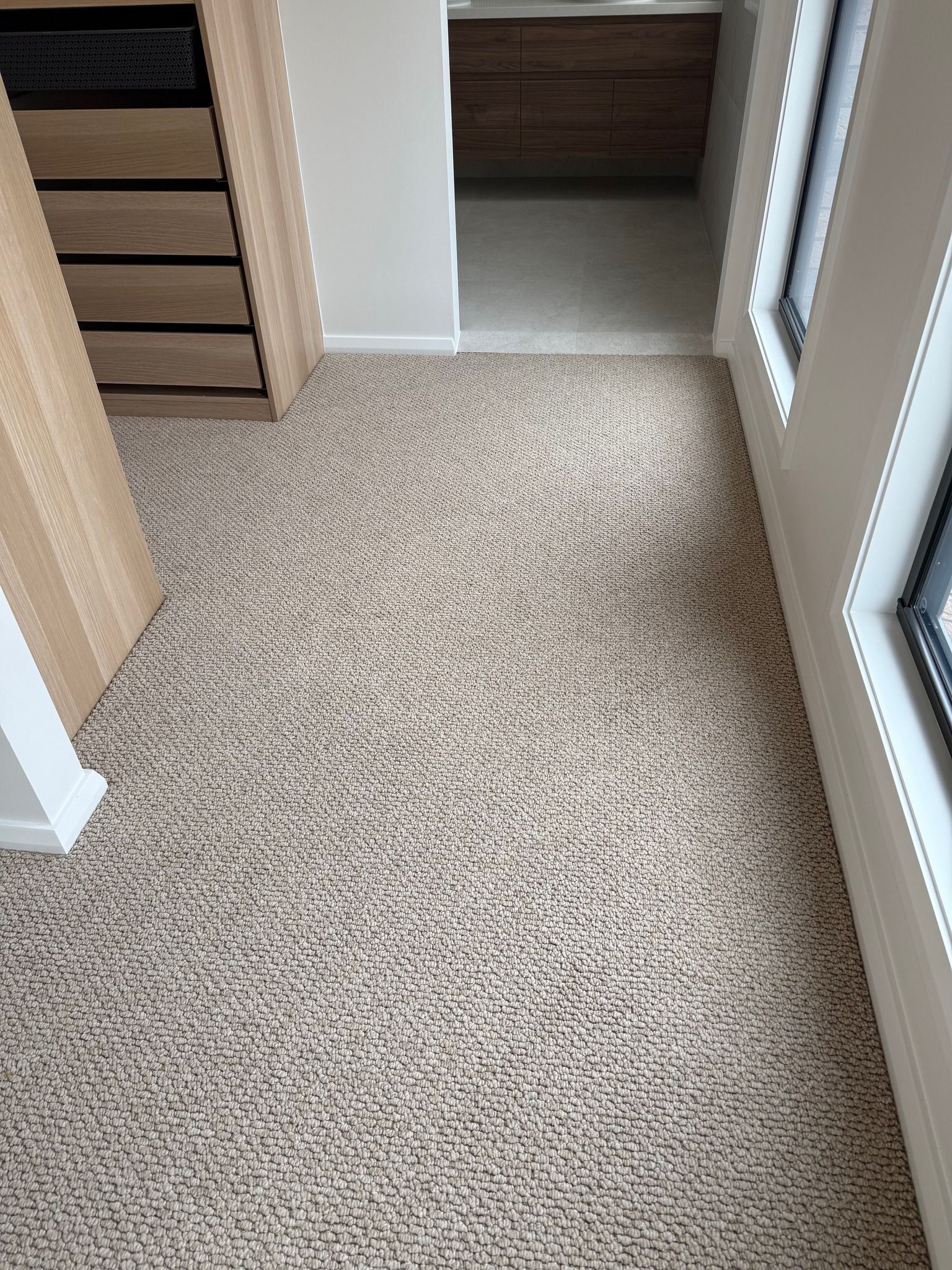 Light brown and beige textured carpet in a room with a wooden wardrobe, white walls, and a window.
