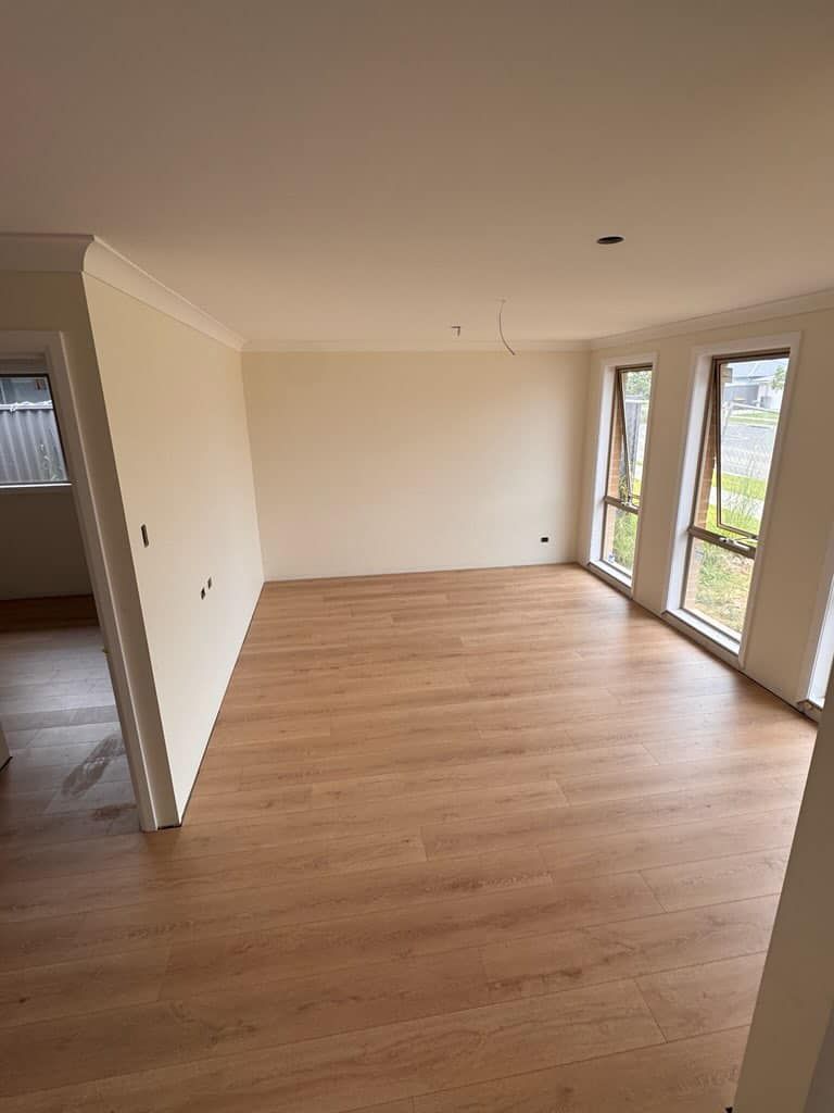 An empty room with light wood-look flooring, cream-colored walls, and two large windows letting in natural light.