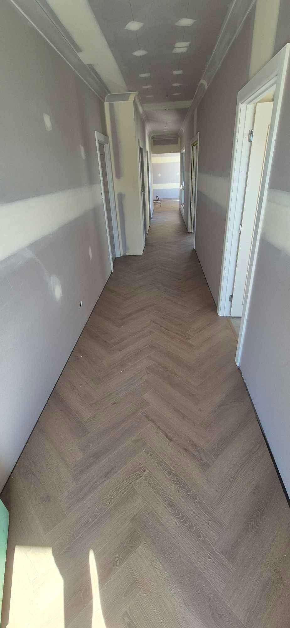 A narrow hallway under renovation with herringbone-patterned wood flooring and grey walls with unfinished plaster joints.