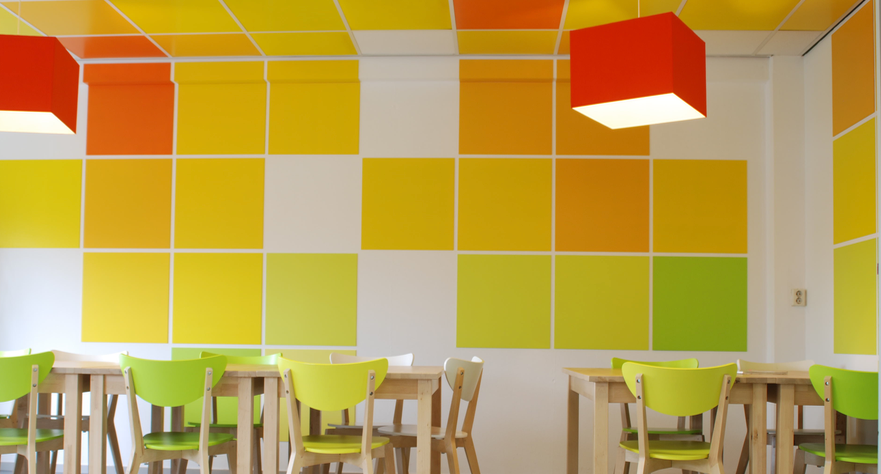 Kleurrijk interieur met gele, oranje en groene accenten, vierkante patronen op de muren en het plafond en groene stoelen.