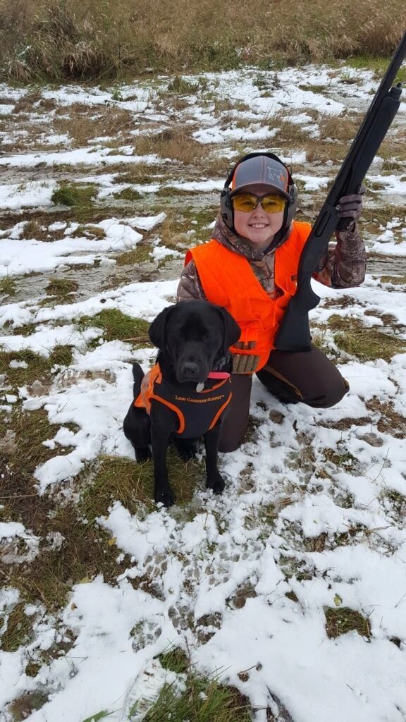 Person and black lab dog in hunting gear kneeling in snowy field, holding shotgun.