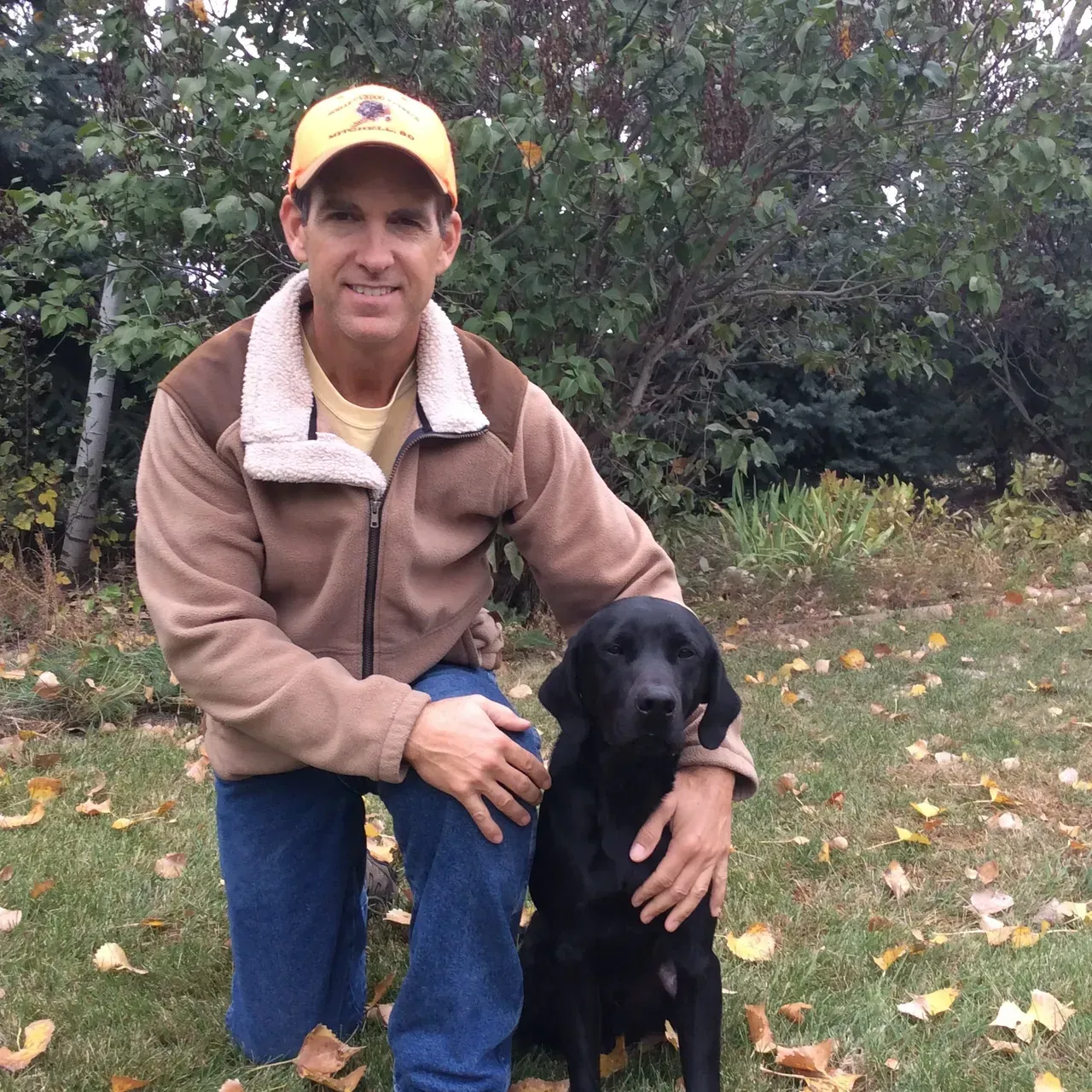 Man kneels with a black Labrador. He wears a brown jacket and orange hat, outdoors on a grassy lawn.