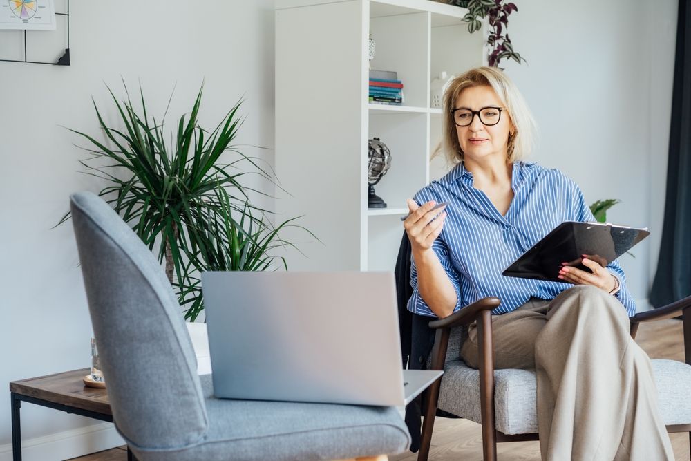 Woman seated indoors holding a tablet, with a laptop on a chair and a potted plant nearby.