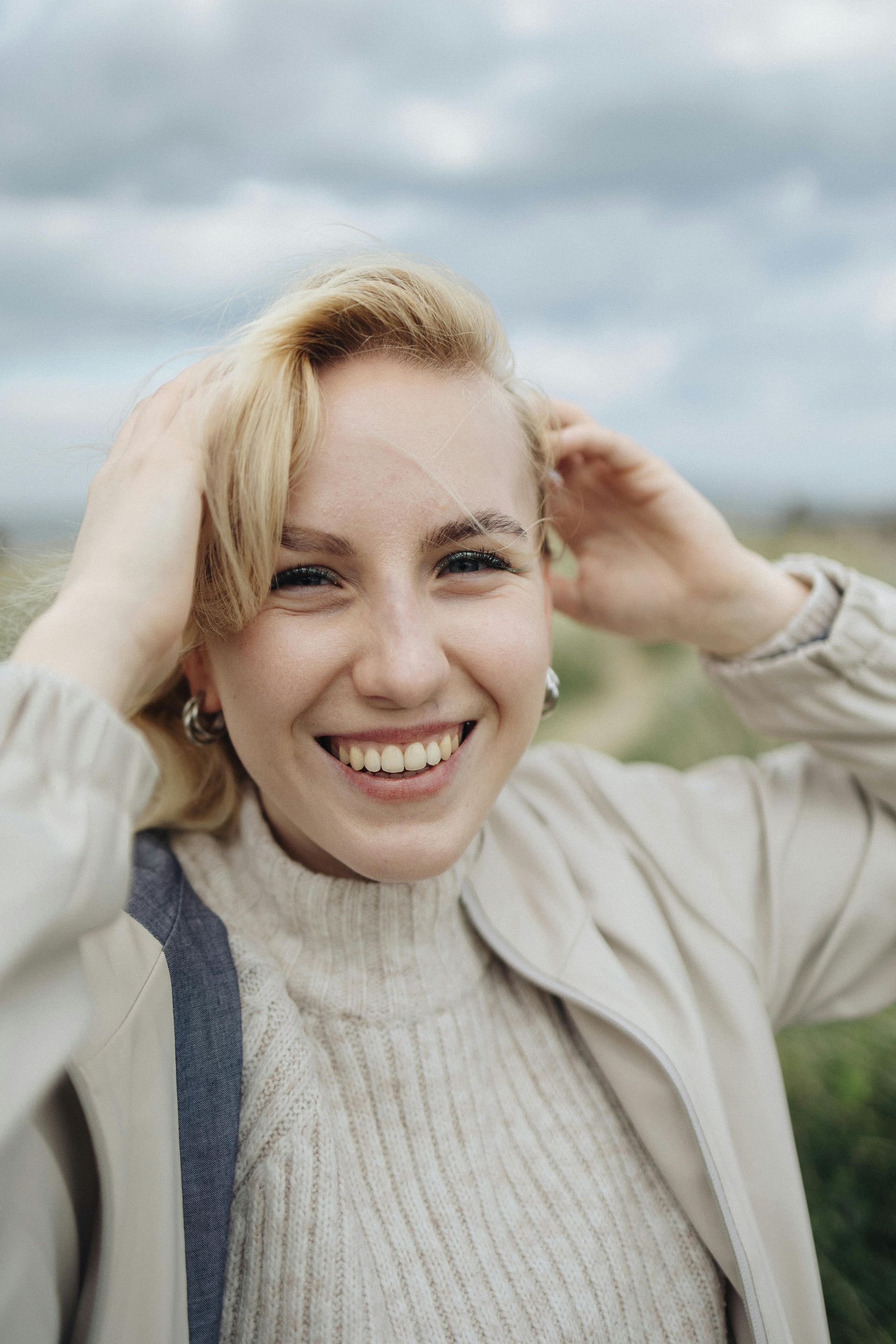 Smiling blonde woman outdoors, wearing a beige sweater, with cloudy sky behind her.