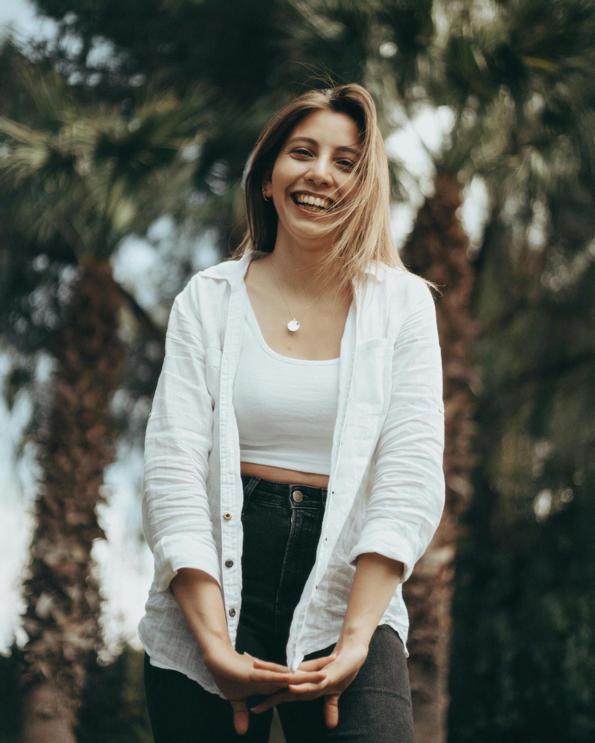 Smiling woman in a white cardigan and black pants standing outdoors with blurred palm trees behind her