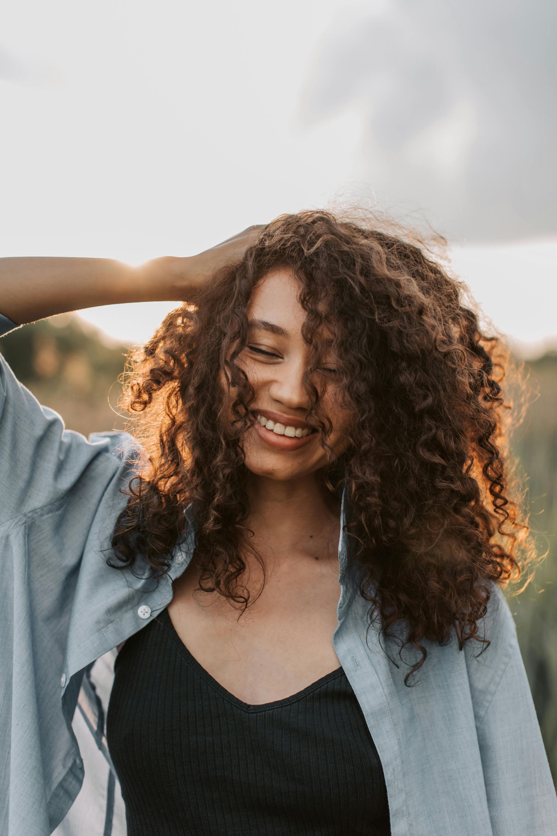 Smiling woman with curly hair standing outdoors at sunset, hand in hair, wearing a black top and light denim shirt