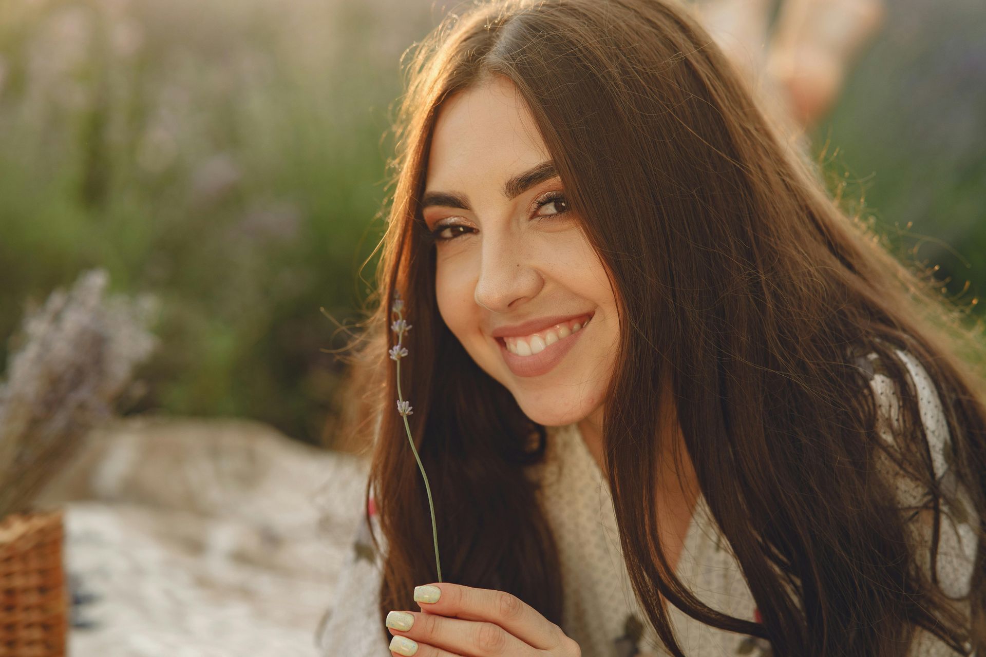 Smiling woman with long brown hair holding a dried flower outdoors in soft sunlight