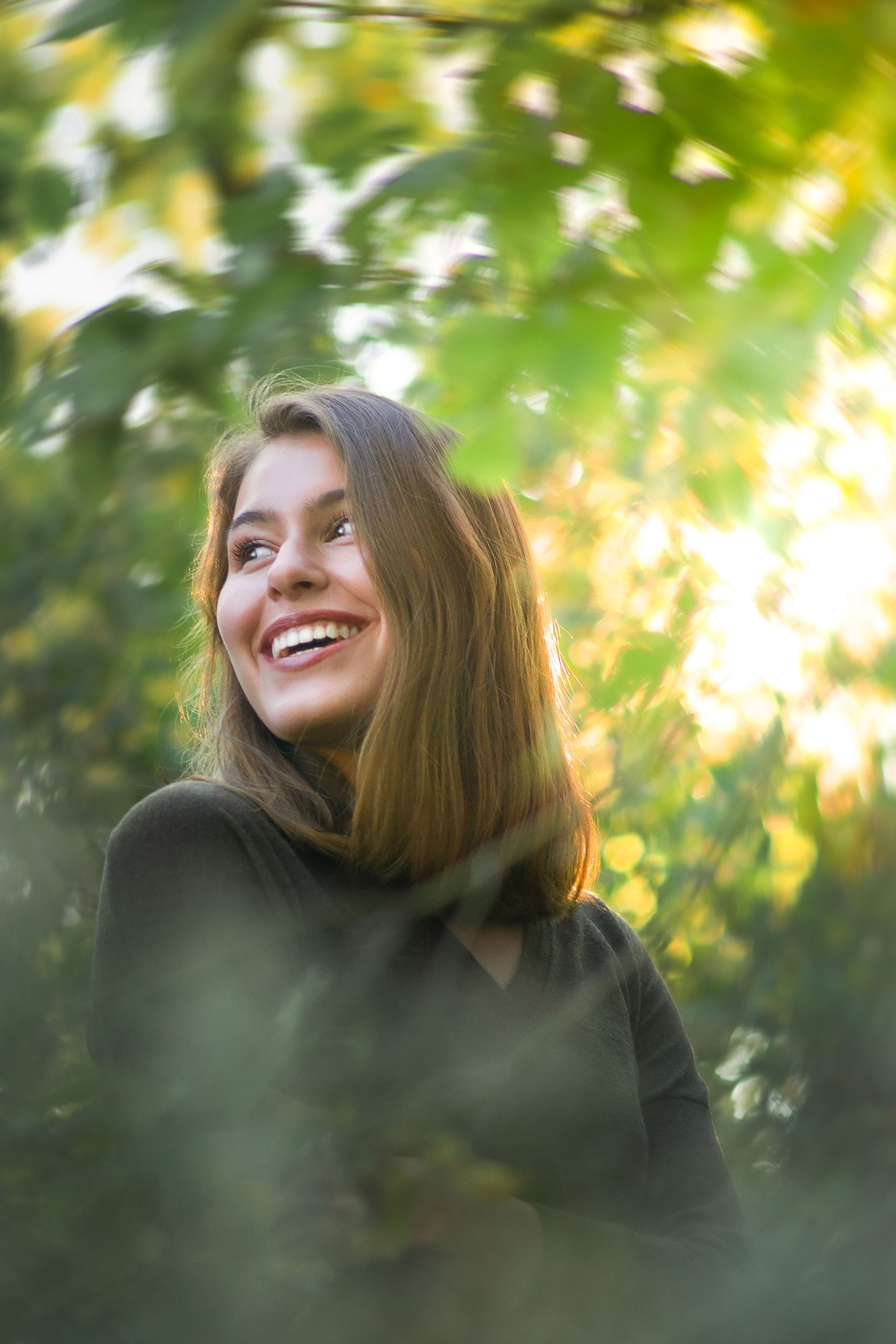 Smiling person in a dark sweater standing among sunlit green leaves outdoors
