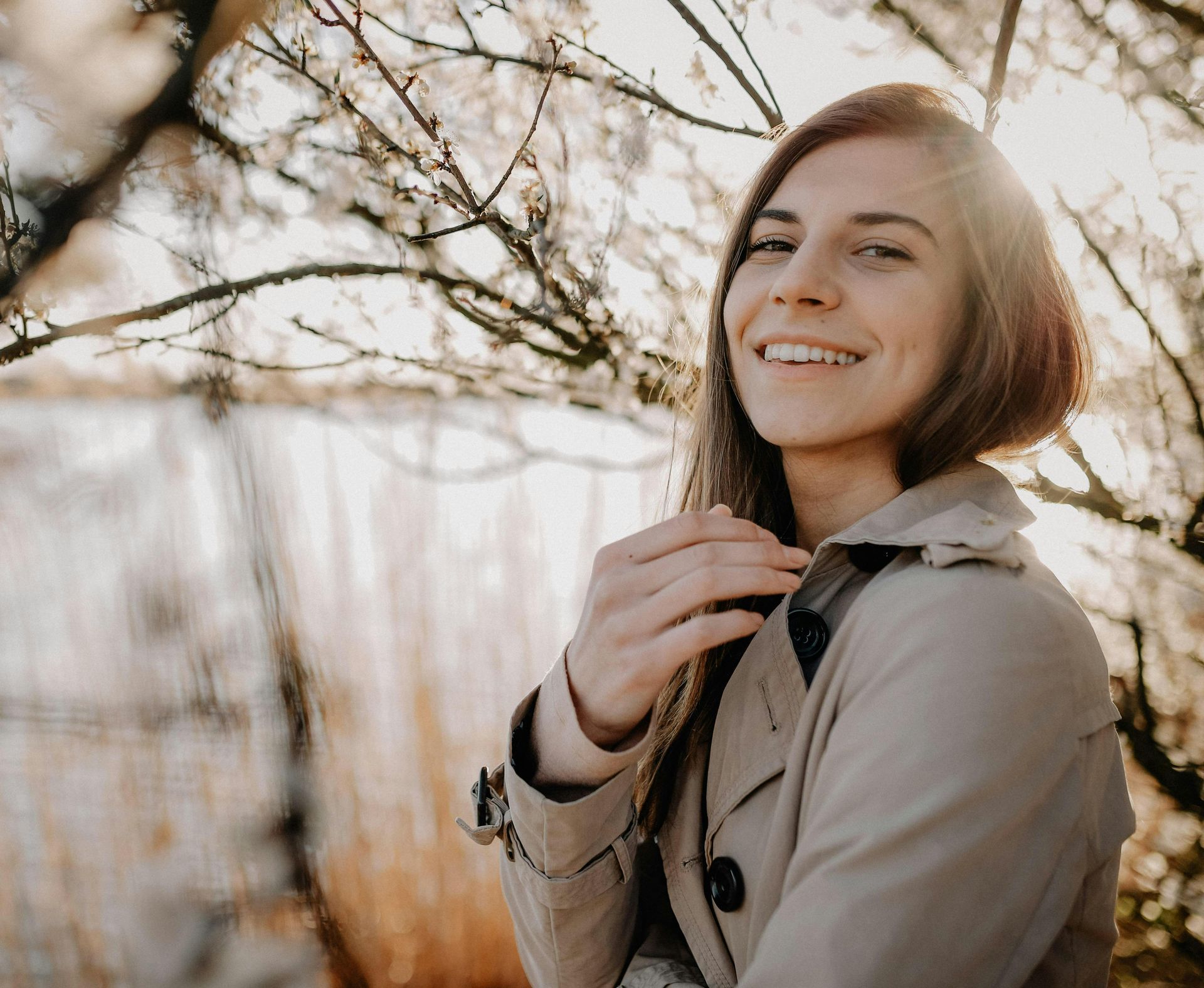 Smiling person in a beige coat standing outdoors by bare branches and tall grass