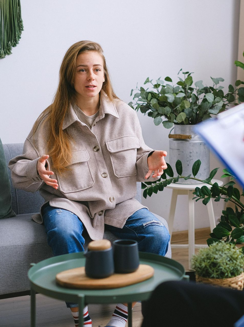Woman speaking in a bright living room with plants, seated on a sofa beside a small table.