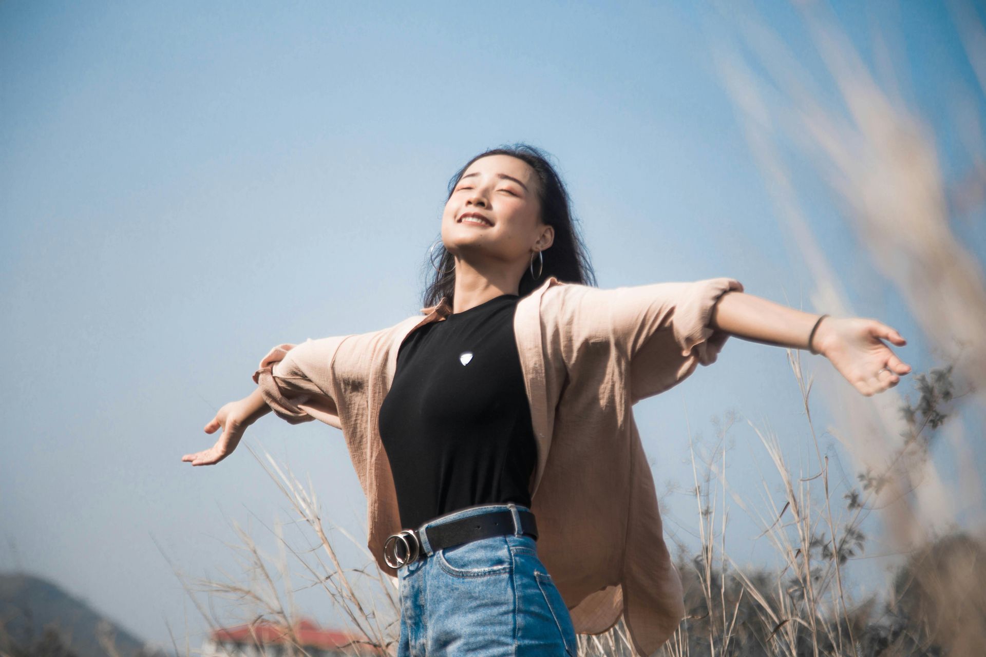 Person in a black top and beige cardigan stands with arms outstretched in a grassy field under a blue sky.