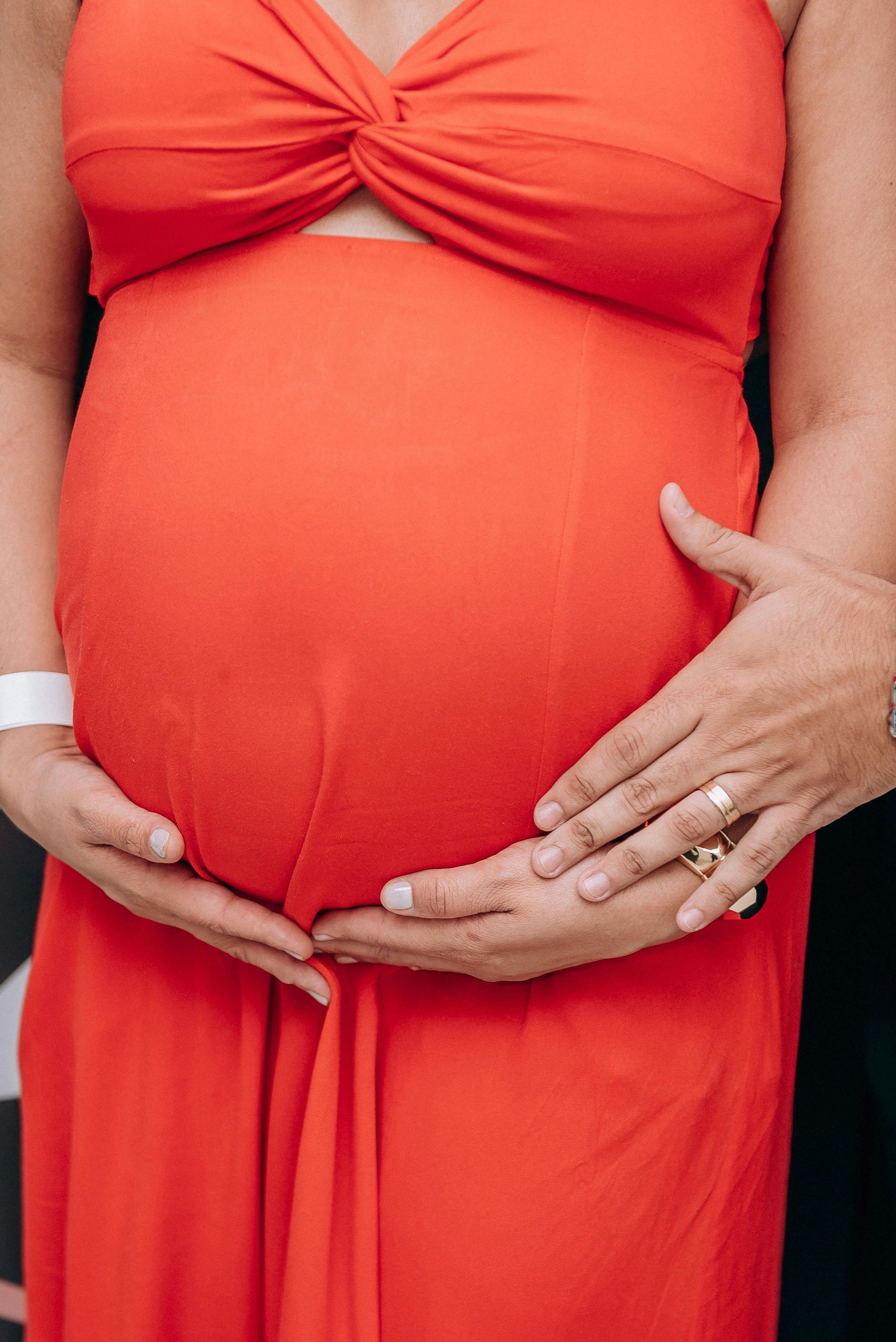 Pregnant woman in red dress, belly held by hands, smiling.