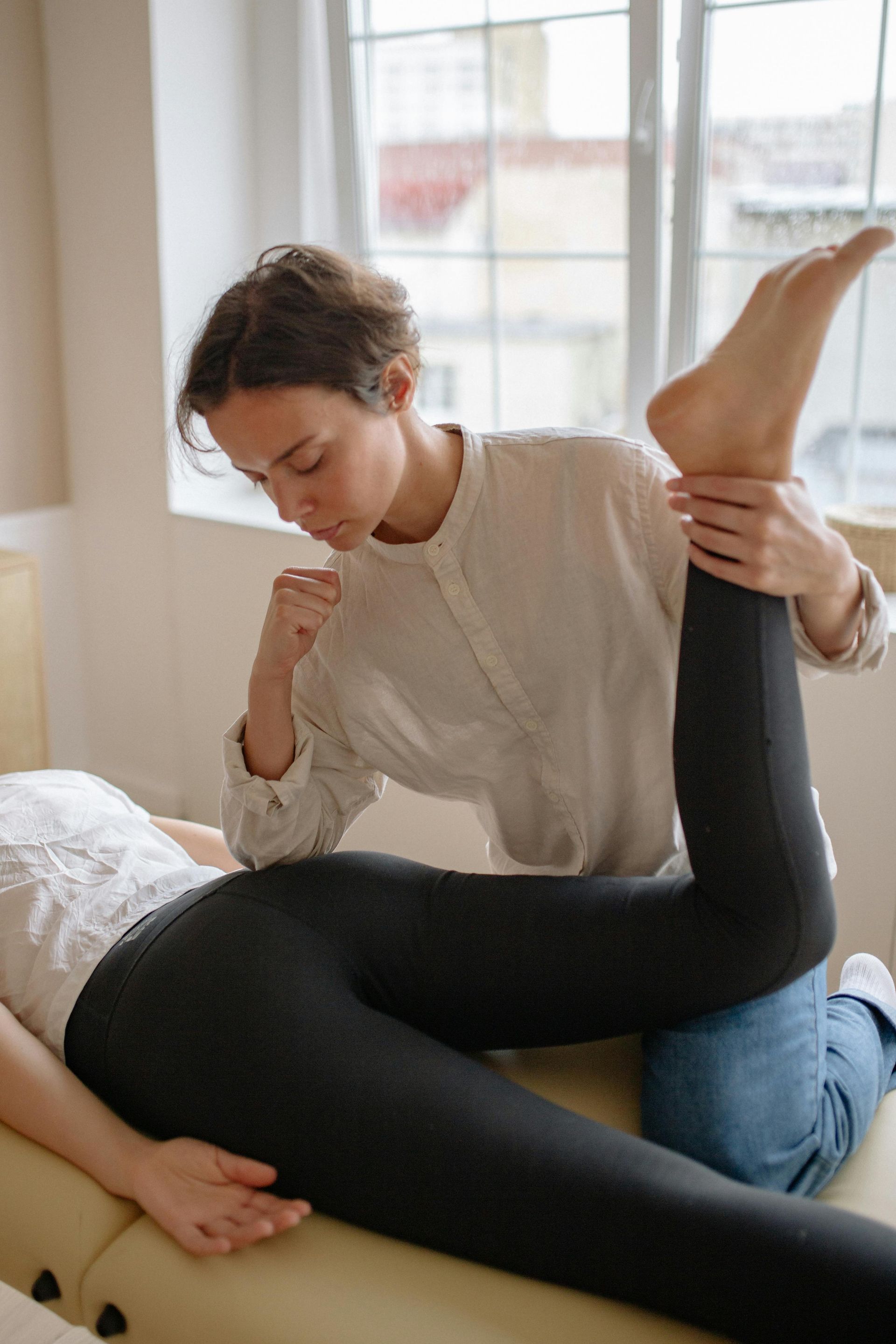 Woman in white shirt stretches another person's leg on a massage table near a window.
