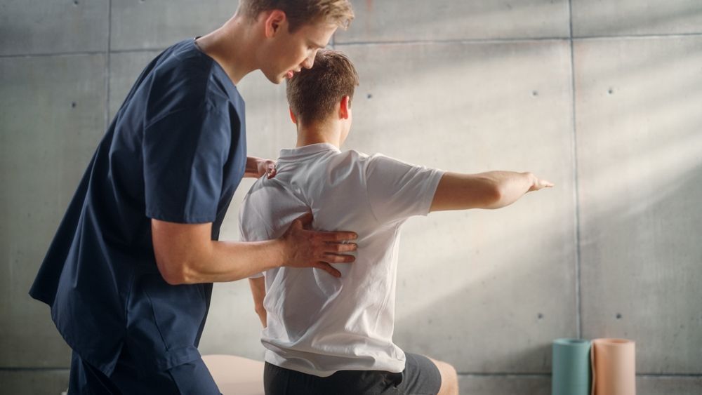 Physiotherapist assisting a patient with spinal mobility. They are in a light-filled room, with neutral colors.