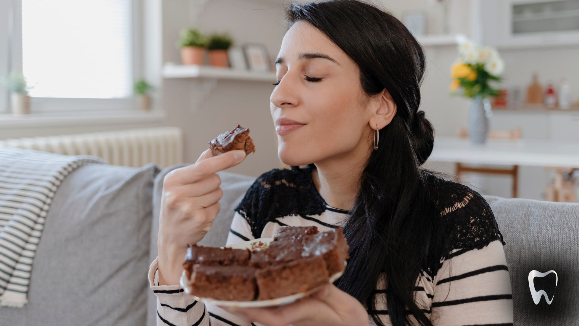Woman enjoying brownie on a couch, smelling it. Indoors with plants and flowers in the background.