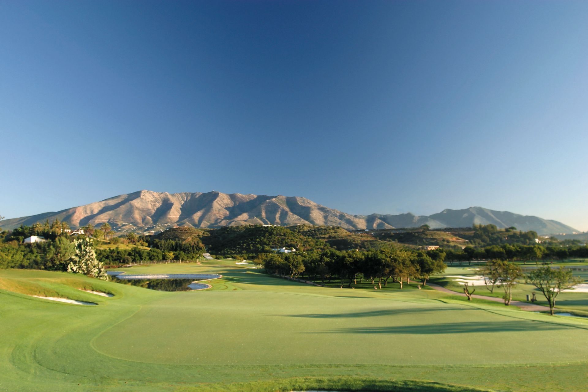 Parcours de golf verdoyant avec des montagnes en arrière-plan sous un ciel bleu clair.