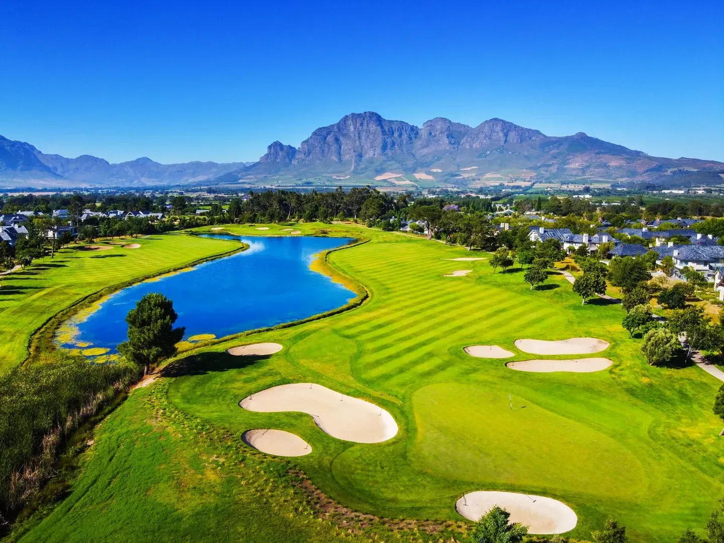 Terrain de golf avec un lac bleu, des bunkers, de l'herbe verte et des montagnes sous un ciel bleu.