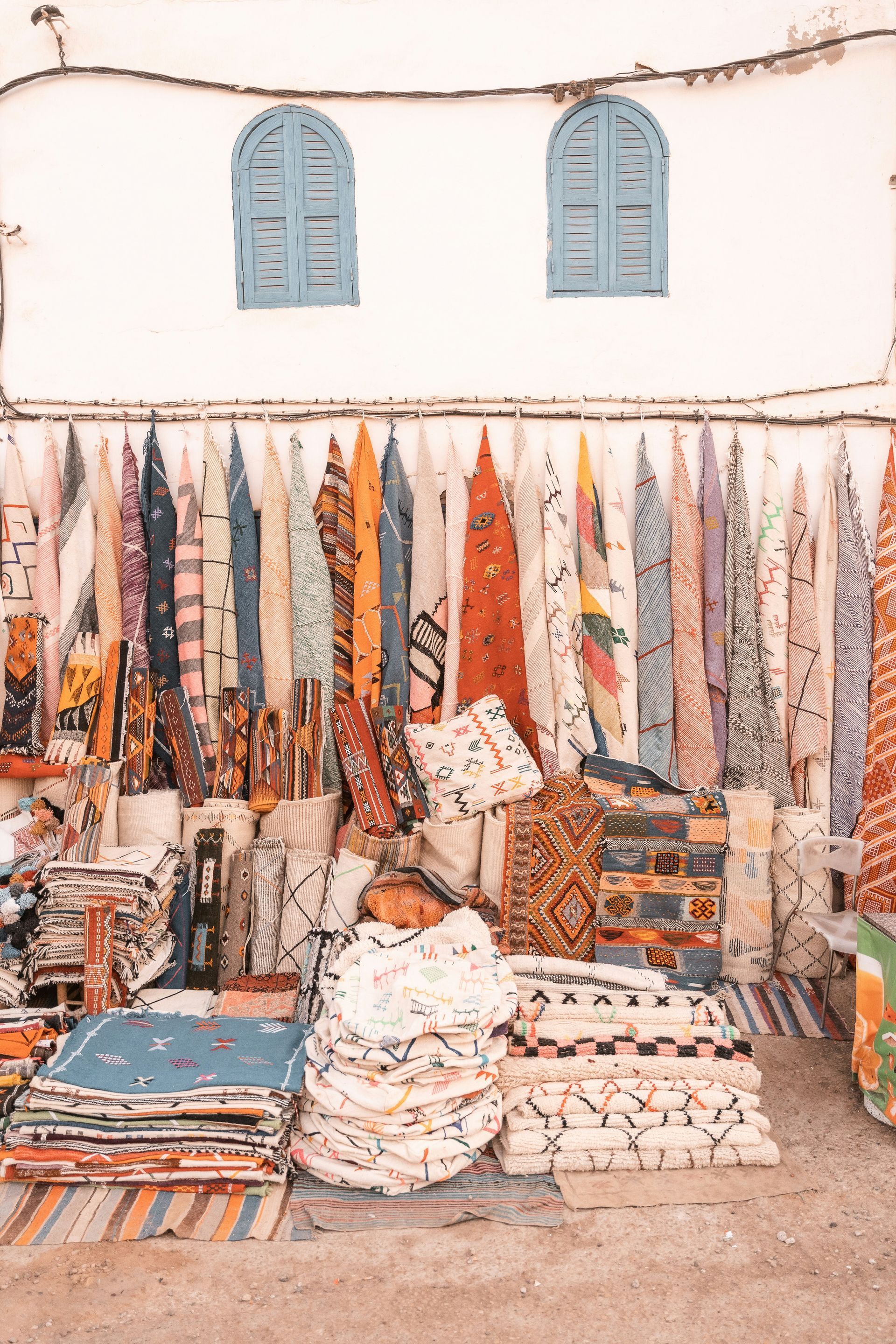 Étal de marché textile coloré avec des tissus suspendus et des tapis pliés devant un mur blanc et des fenêtres bleues