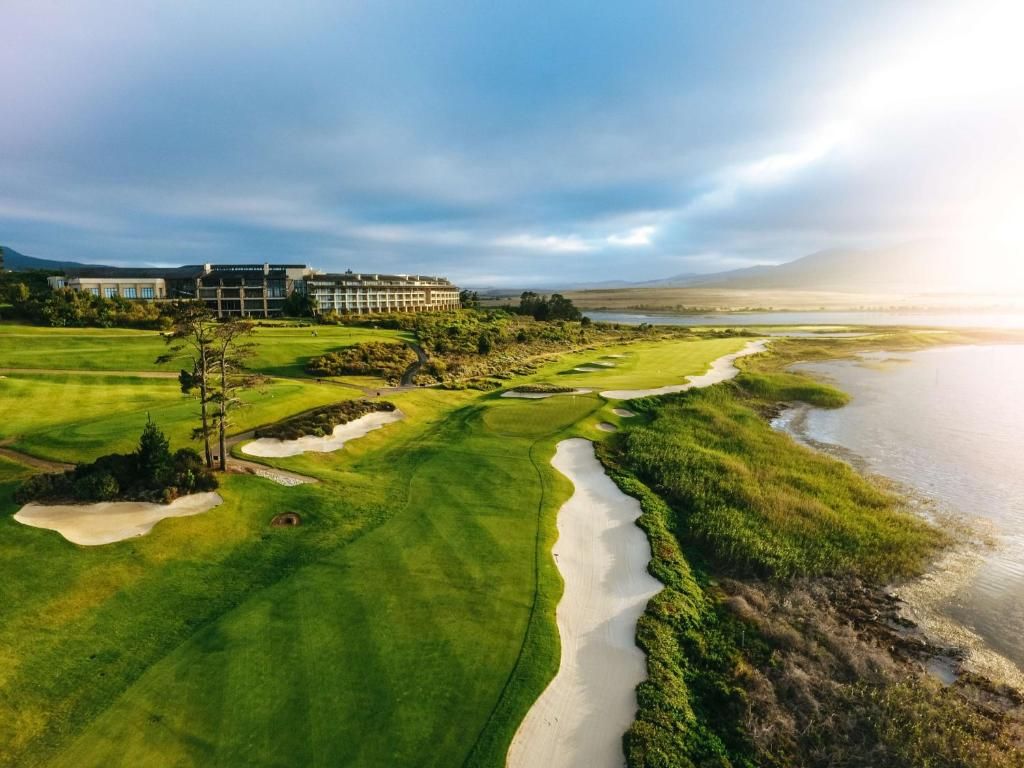 Vue aérienne d'un terrain de golf avec un bâtiment en arrière-plan, près d'un plan d'eau, sous un ciel nuageux.