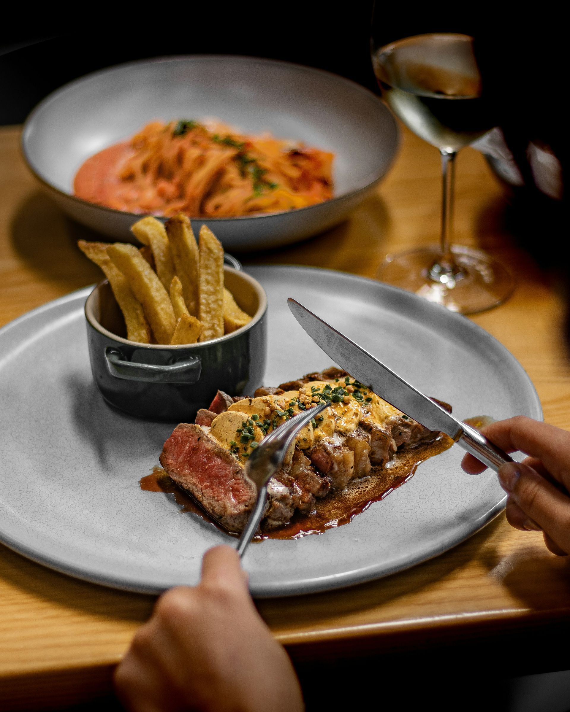 Des mains découpant un steak saisi à la poêle sur une table de restaurant, avec des frites et des pâtes en arrière-plan