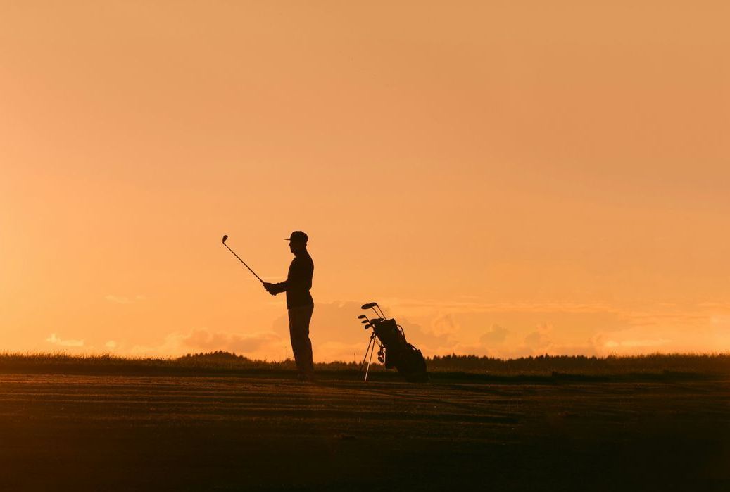 Silhouette d'un golfeur et de son chien au coucher du soleil sur un champ, sous un ciel orangé.