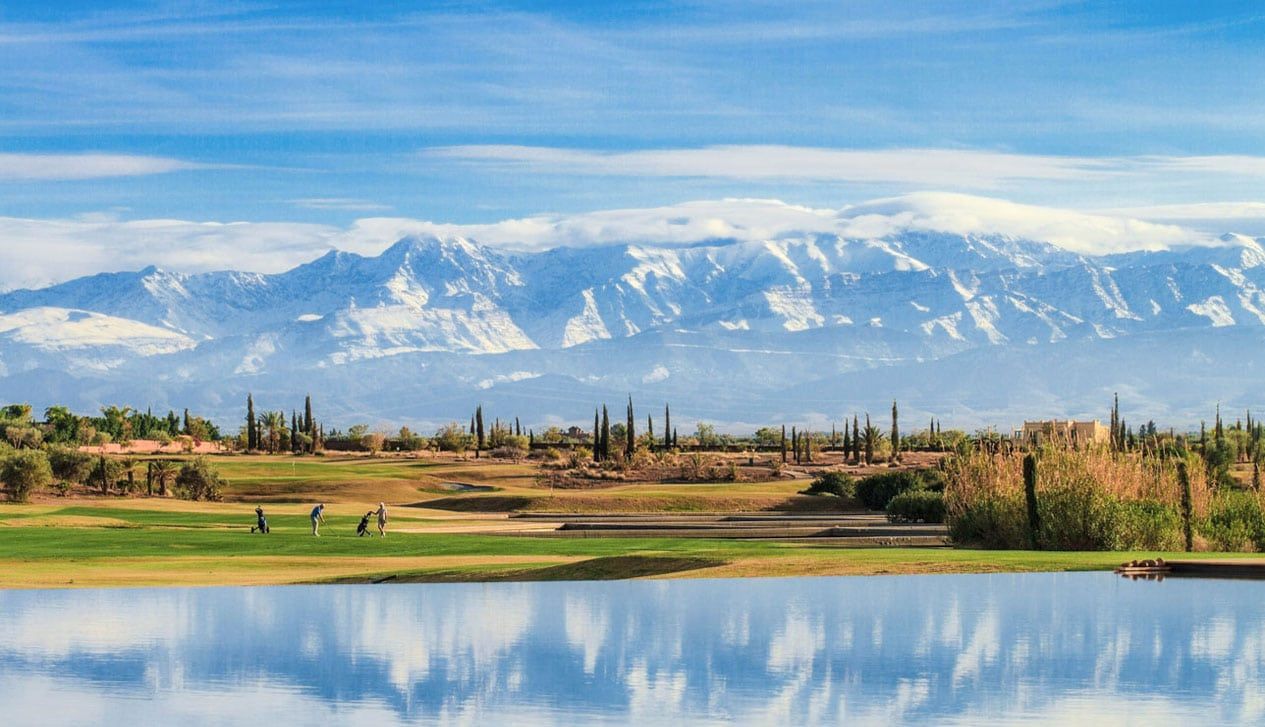 Terrain de golf avec plan d'eau, paysage désertique et montagnes enneigées sous un ciel bleu.