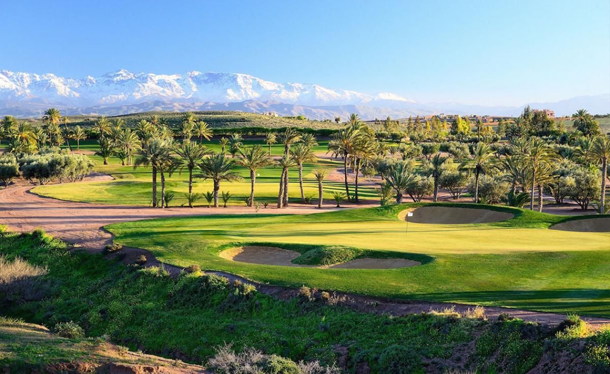 Parcours de golf ensoleillé avec fairways verdoyants, bunkers de sable, palmiers et collines au loin sous un ciel bleu.