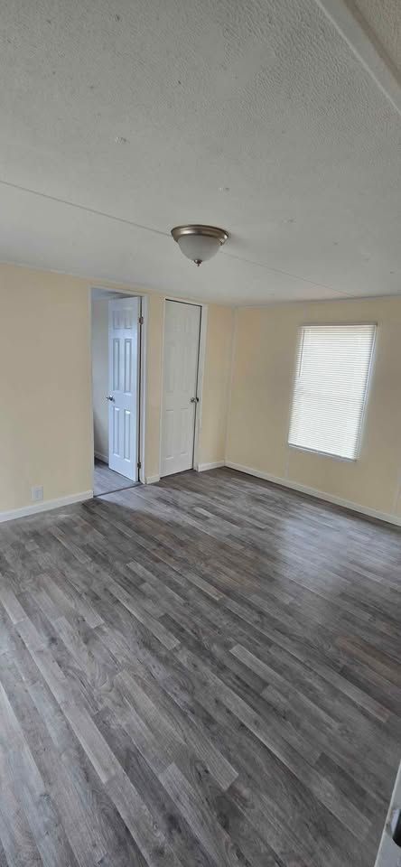 Empty room with gray wood floors, beige walls, a ceiling light, and a window with blinds.