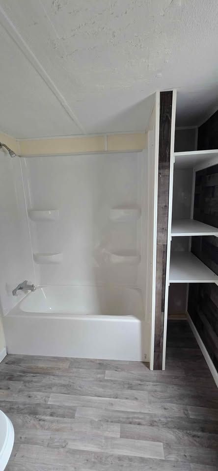 Small bathroom with a white tub, tiled shower walls, and built-in shelving beside gray wood-look flooring.