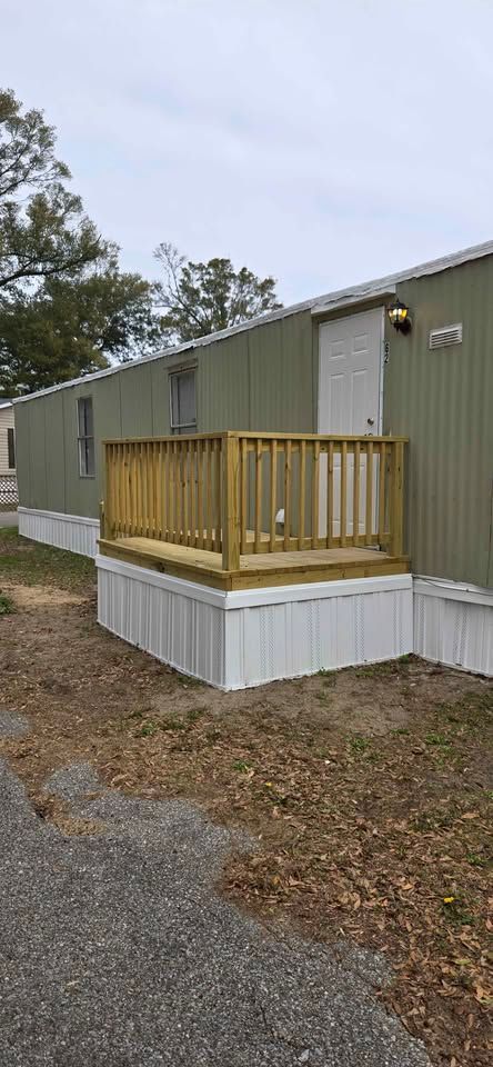 Green mobile home with small wooden porch and white skirting beside a gravel drive