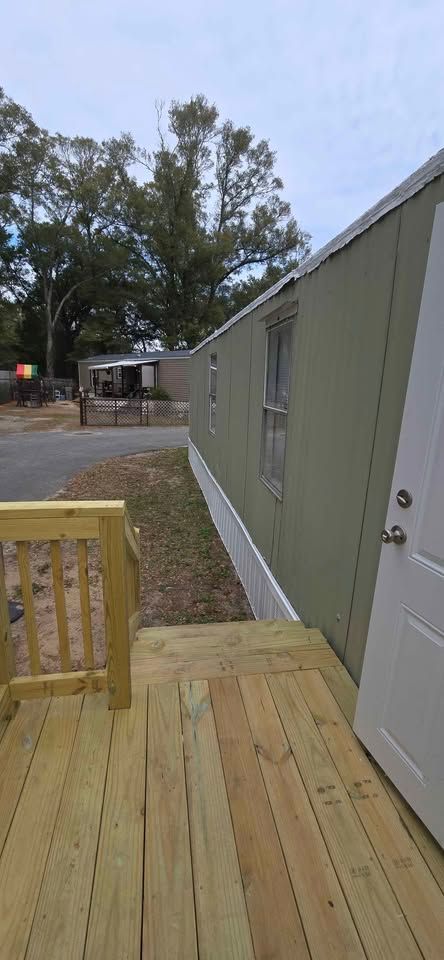 Wooden porch beside a gray mobile home, with a dirt yard and trees in the background.