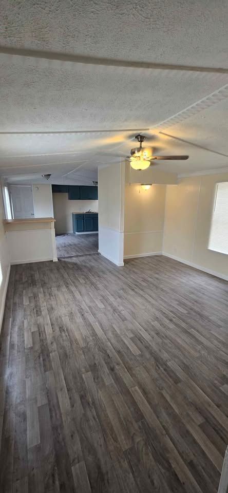 Empty living room with gray wood floors, white walls, ceiling fan, and kitchen area in the back.