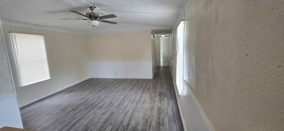Empty living room with gray wood floors, white walls, ceiling fan, and a window with blinds.