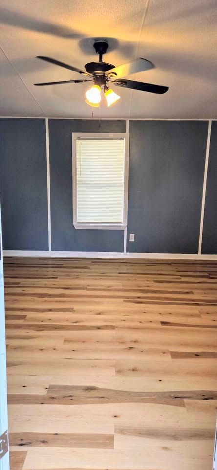 Empty room with gray walls, white trim, wood floor, ceiling fan, and a single window with blinds.