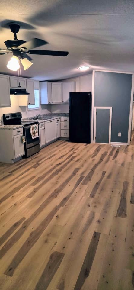 Empty kitchen and living area with wood floors, white cabinets, dark walls, and ceiling fan