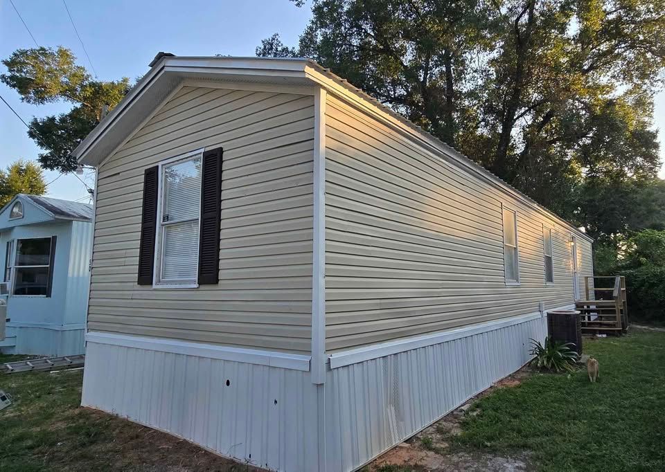 Single-wide beige mobile home with white skirting and dark shutters in a grassy yard at dusk