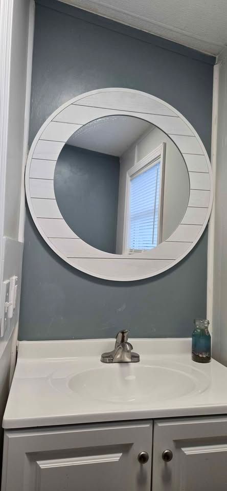 White round mirror above a bathroom sink with gray walls and a small window reflection