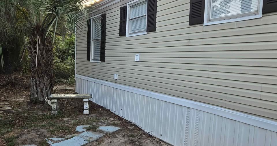 Exterior side of a beige mobile home with white skirting and a small bench beside a stone walkway