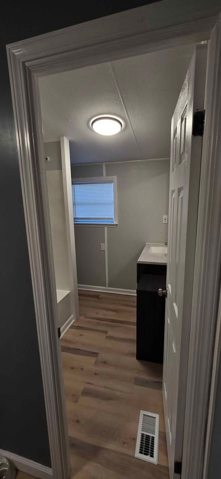 Narrow hallway with white walls, wood floor, ceiling light, window, and a doorway to a small room.