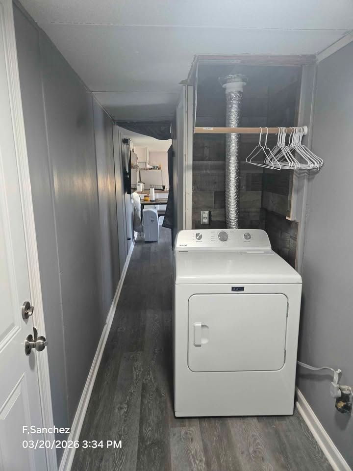 Narrow laundry room hallway with white dryer, hanging rack, and gray walls leading to another room