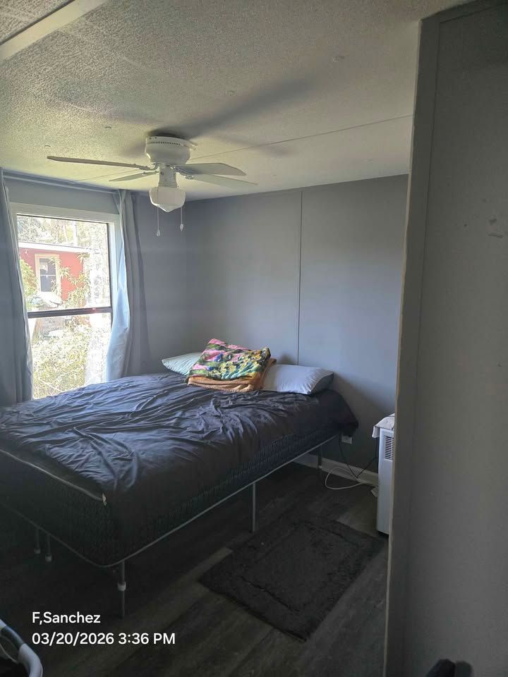 Small bedroom with a made bed, gray walls, ceiling fan, and window letting in daylight.