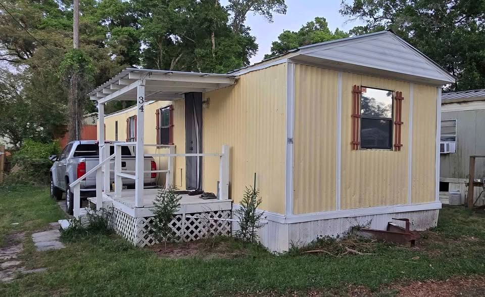 Yellow mobile home with a white porch and gray roof in a grassy yard, parked beside a white truck