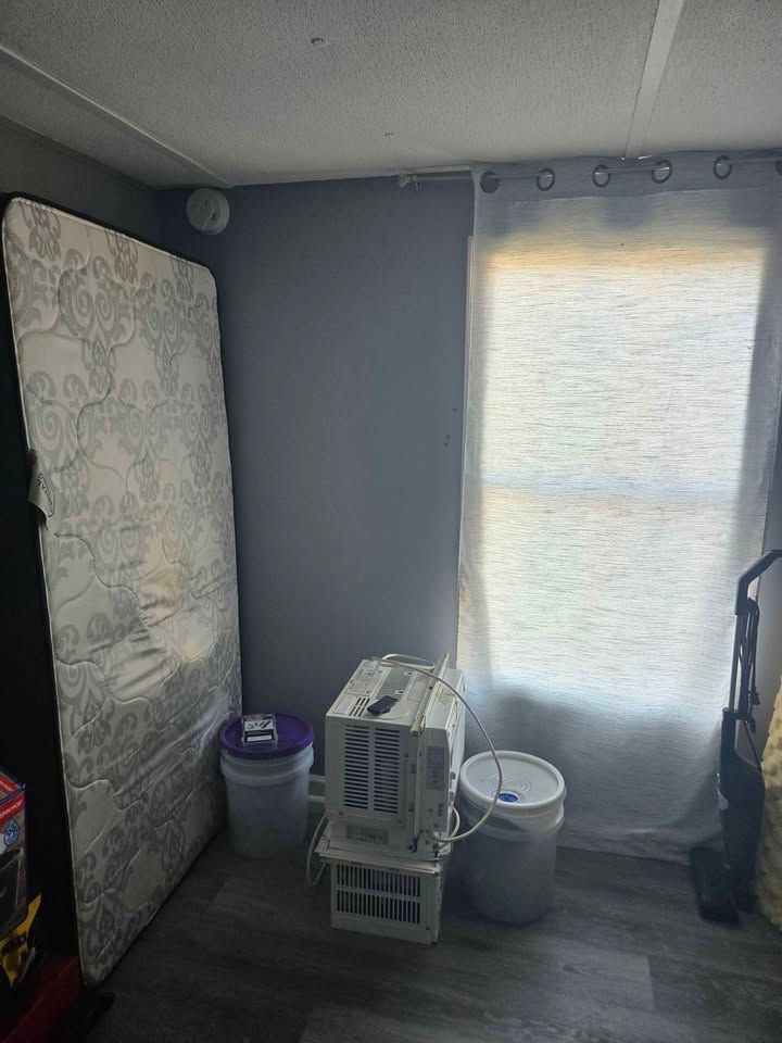 Small room with a mattress, storage bins, and a window covered by a sheer curtain.